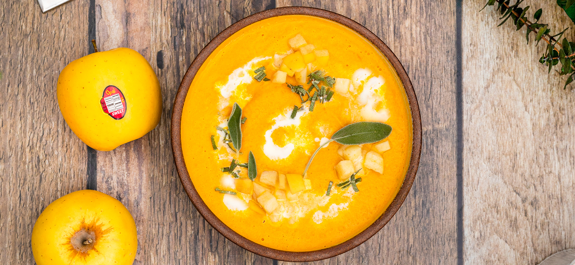 Bright orange carrot and apple soup in the center with swirled cream on top and two yellow Opal apples to the right. 